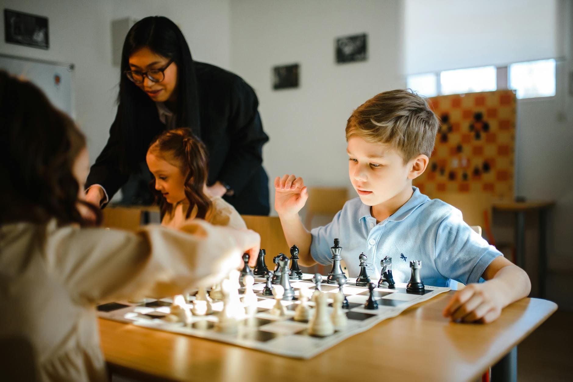 Young children playing chess under adult supervision in a classroom setting.