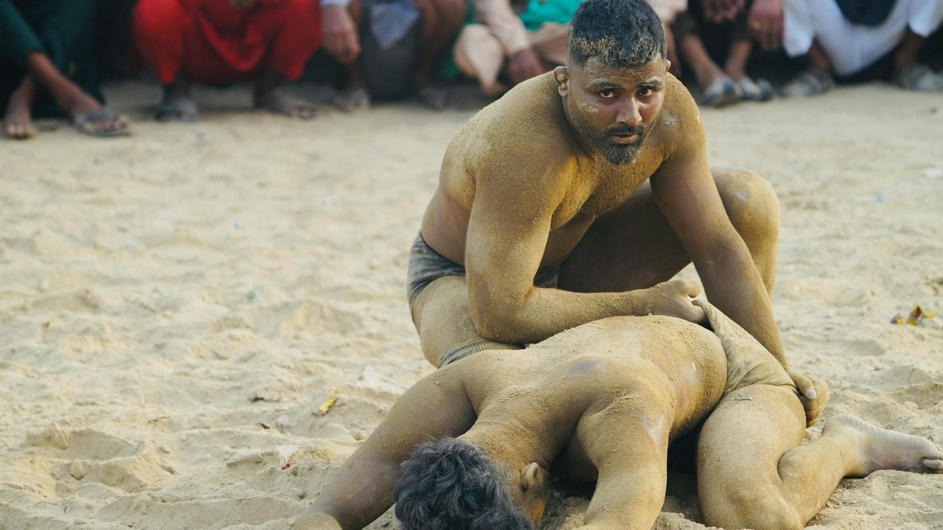 Two men engaged in a traditional mud wrestling match outdoors, surrounded by spectators.
