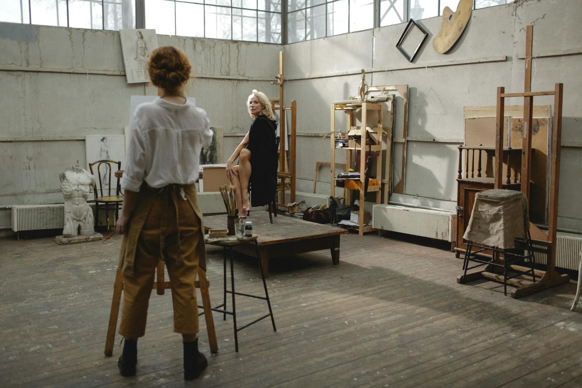Two women engaged in artistic work inside a bright, spacious studio filled with art supplies and easels.