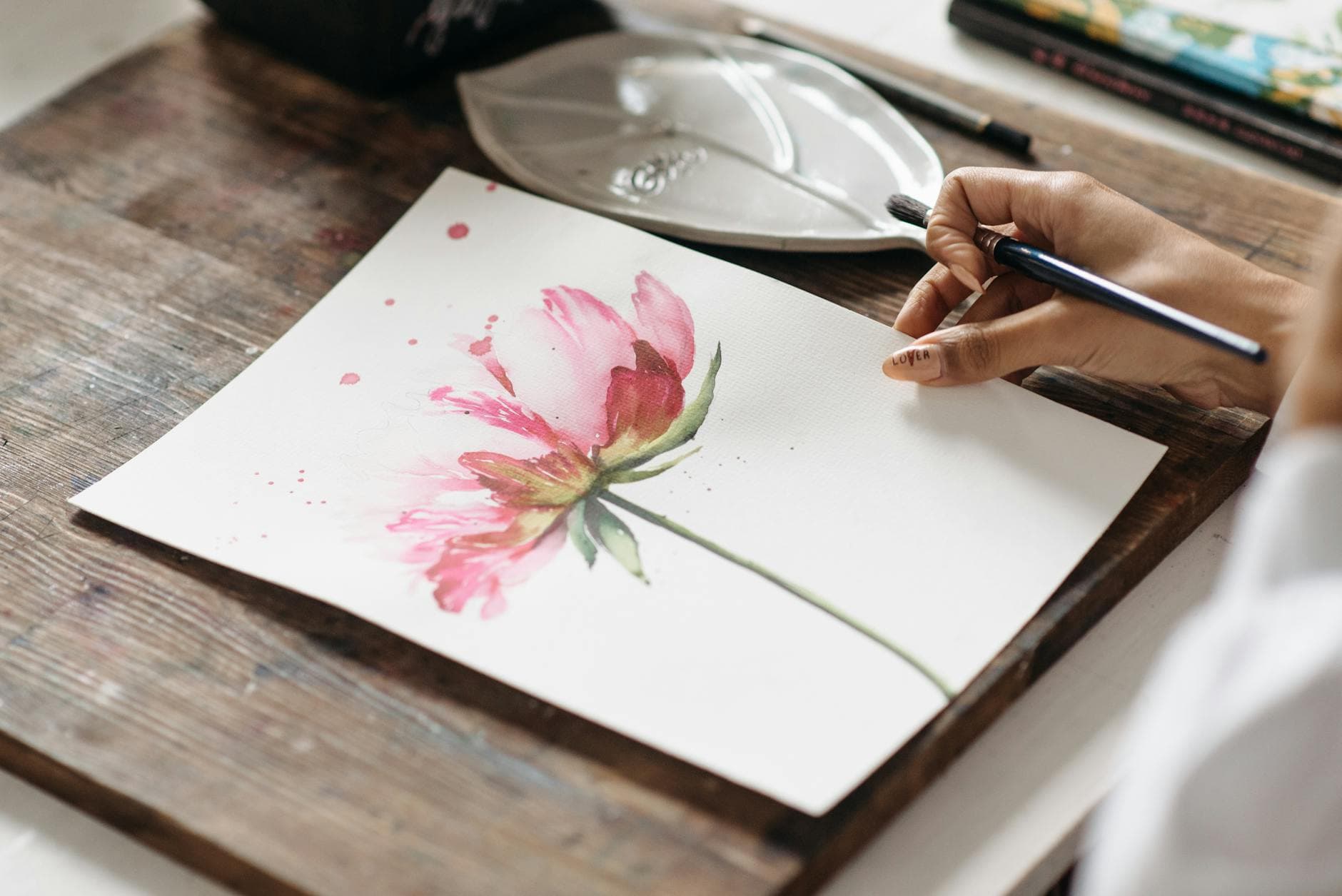 Close-up of a hand painting a pink flower with watercolor on white paper.