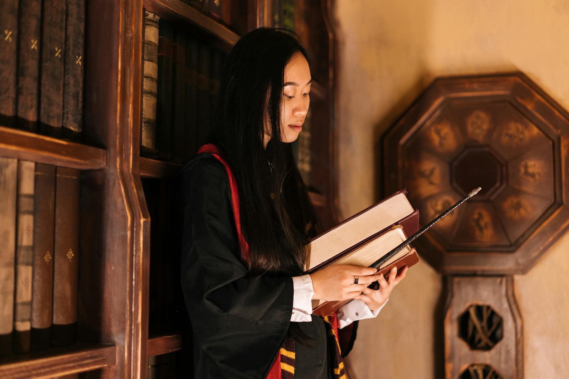 A young woman in a wizard costume studies books with a wand inside a classic library.