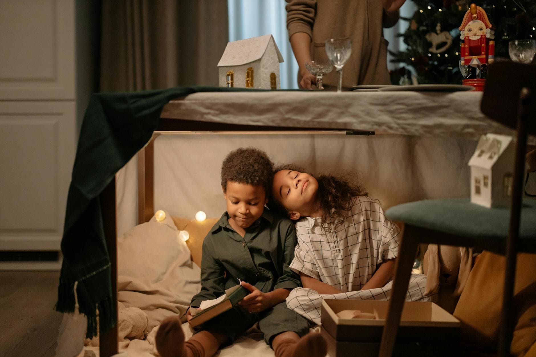 Two children sitting under a table enjoying a Christmas scene with cozy decorations.