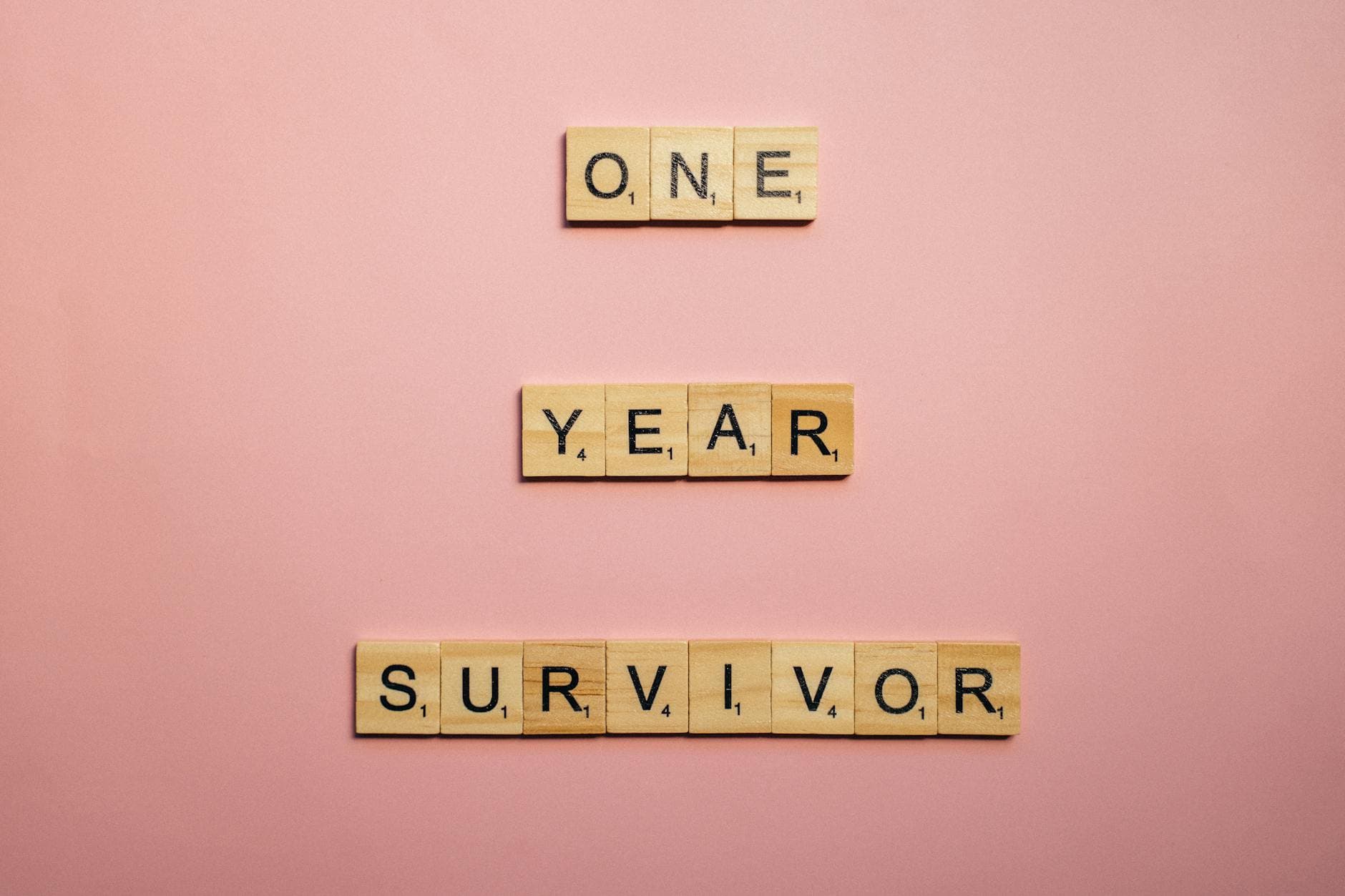 Wooden tiles spell 'One Year Survivor' on a pink background, symbolizing strength and resilience.