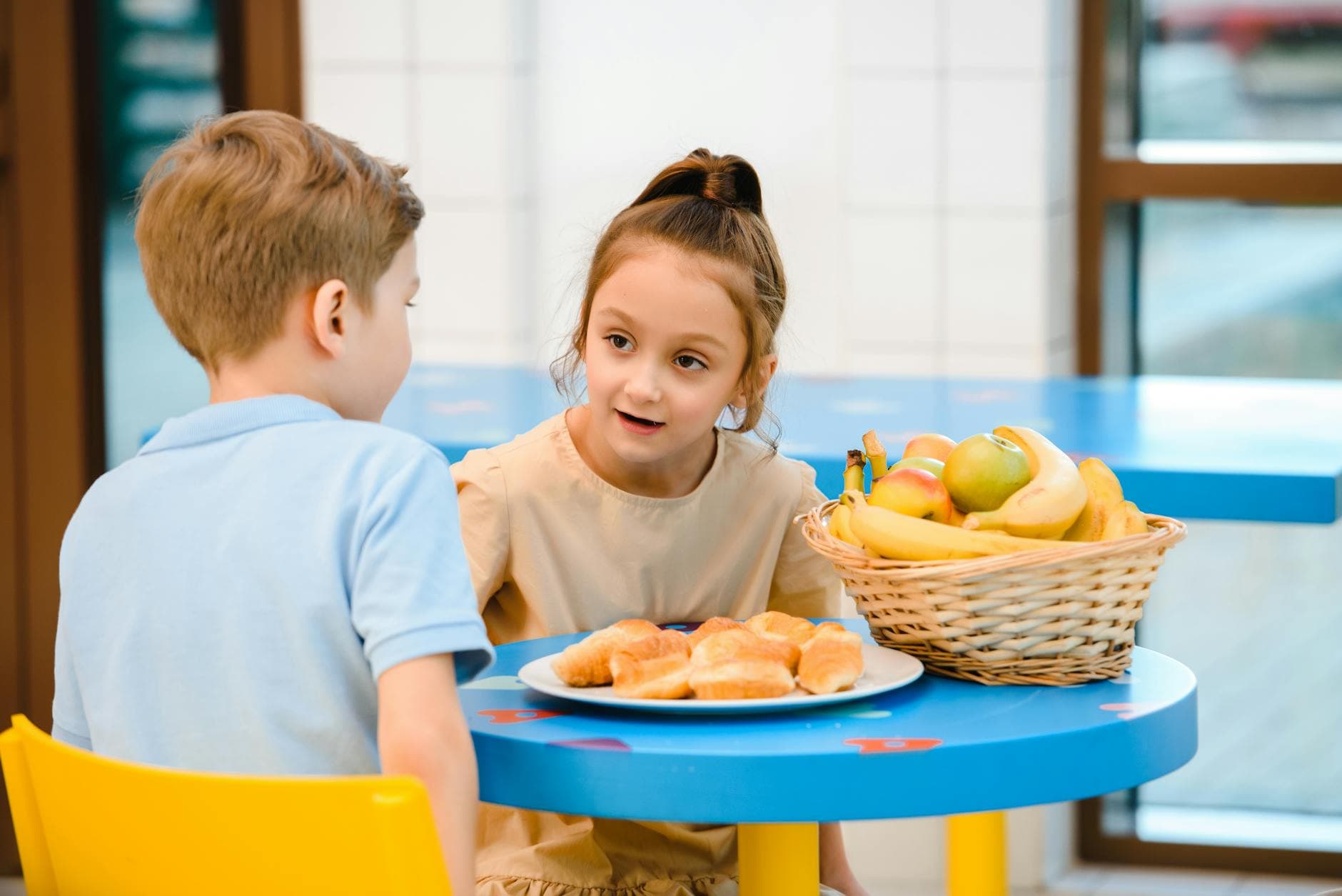 Two children engaging in friendly conversation over breakfast with fruit and pastries.