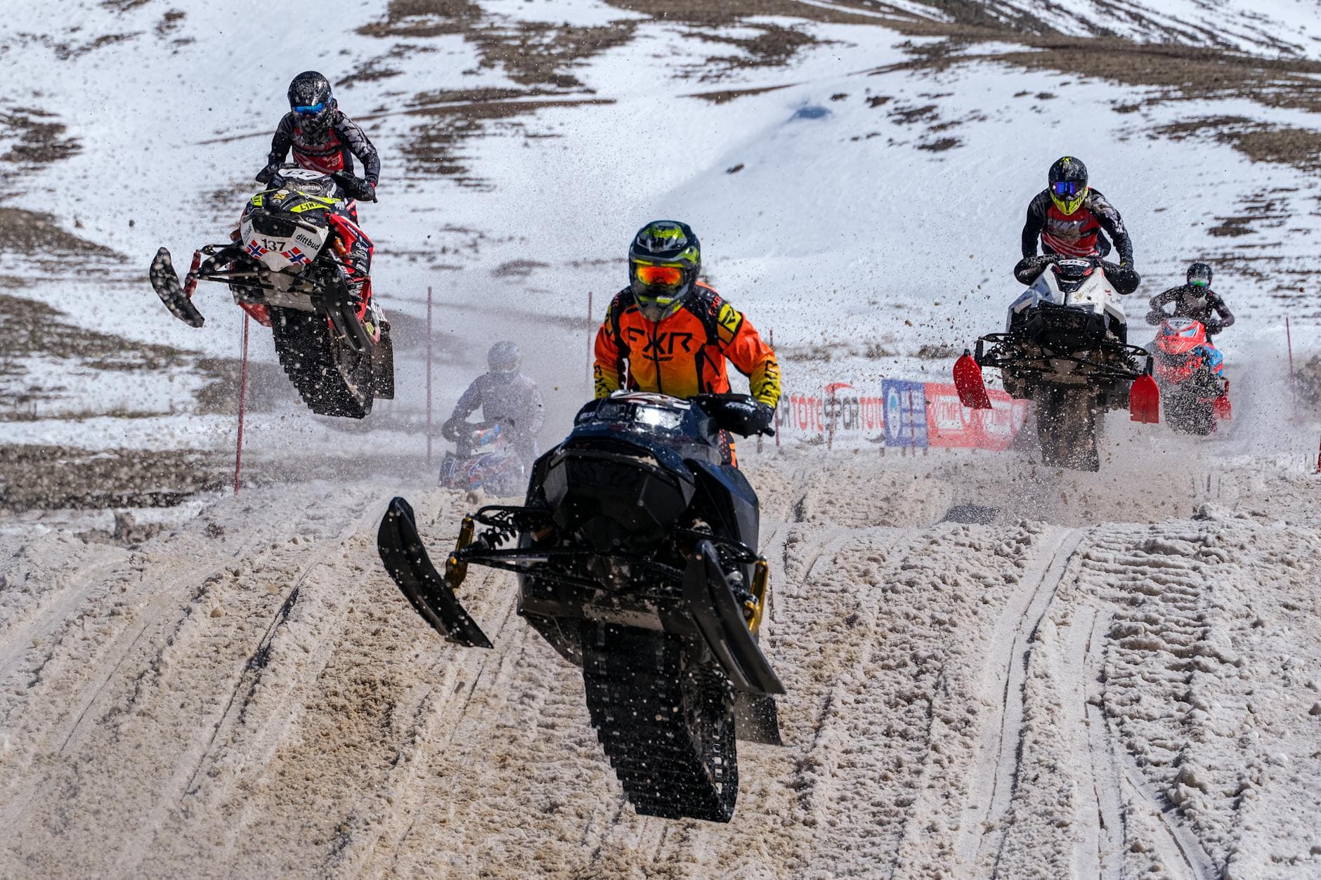 Action-packed snowmobile race with riders launching on snowy track against a mountainous backdrop.