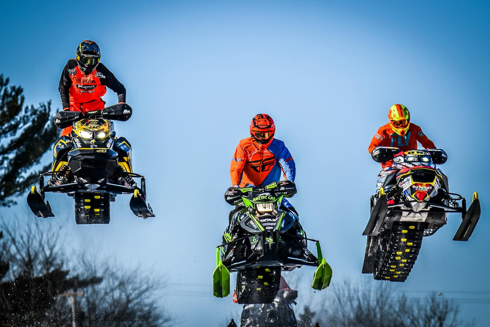 Three snowmobilers perform mid-air jumps during a sunny winter day.