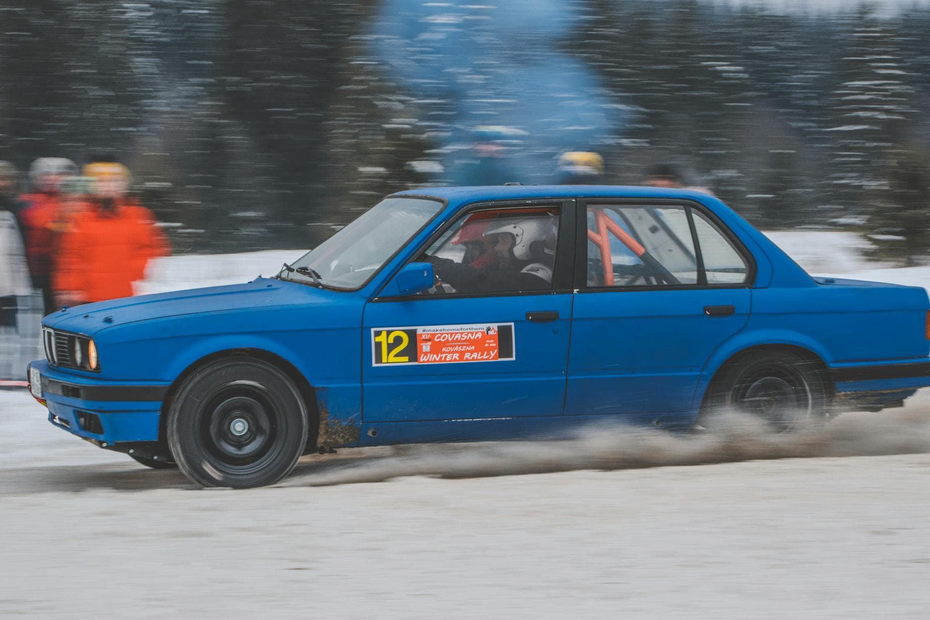 A blue car speeds through snow during the Covasna Winter Rally in Romania, showcasing winter motorsport action.