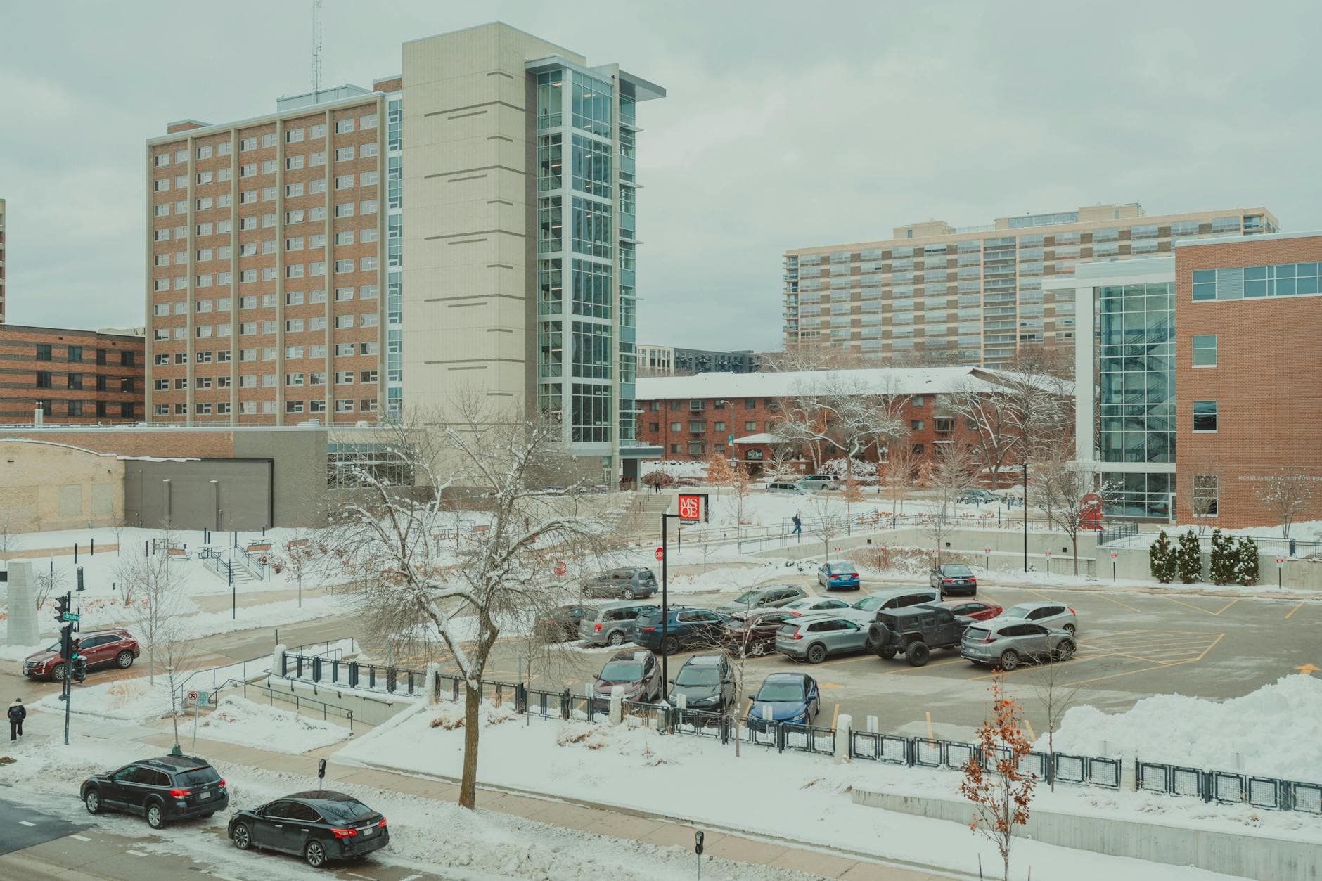 Snow-covered urban scene with Milwaukee campus buildings and parking lot in winter.