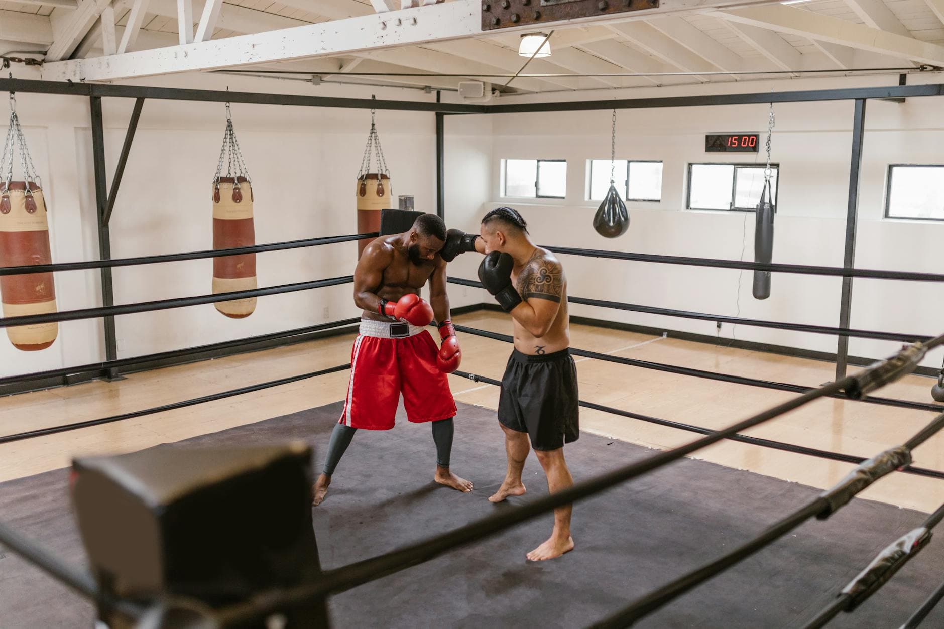 Two men engaged in a sparring session inside a boxing ring at an indoor gym with hanging punching bags.