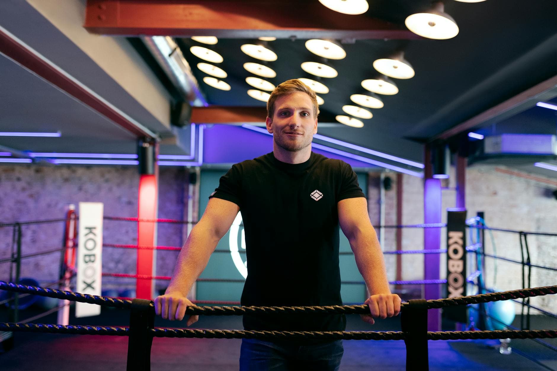 A male trainer stands confidently in a modern boxing gym with vibrant lighting.