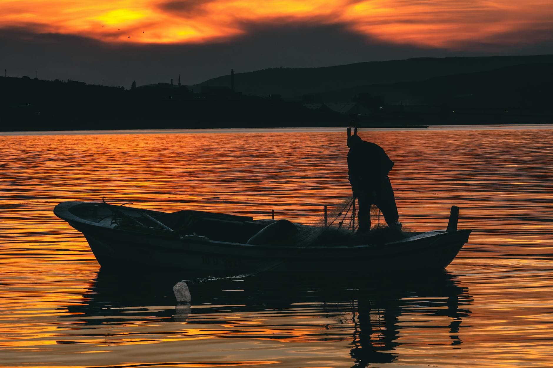 A silhouette of a fisherman casting nets on a small boat during a vibrant orange sunset on a calm lake.