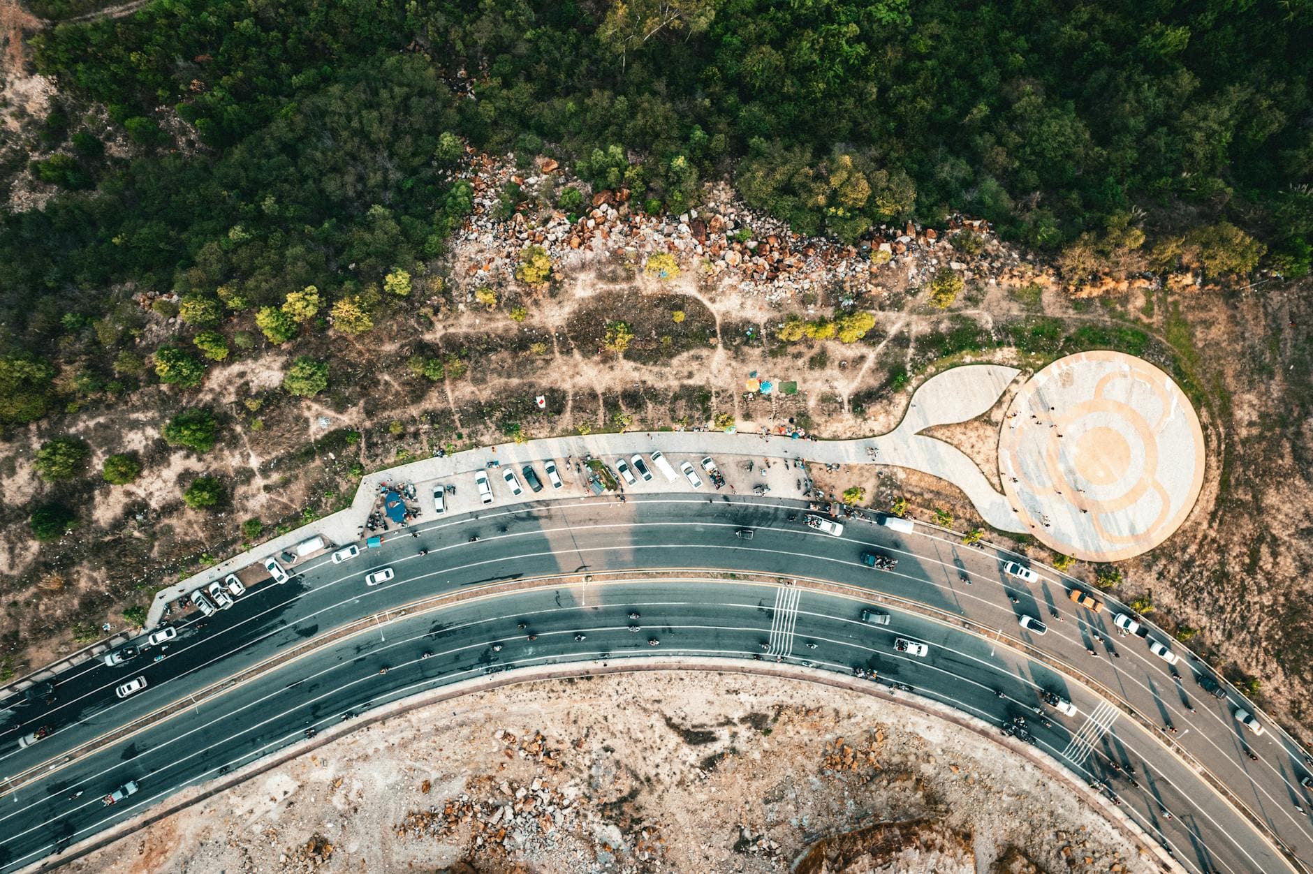 Drone shot of a curving highway bordered by parked cars and lush greenery.