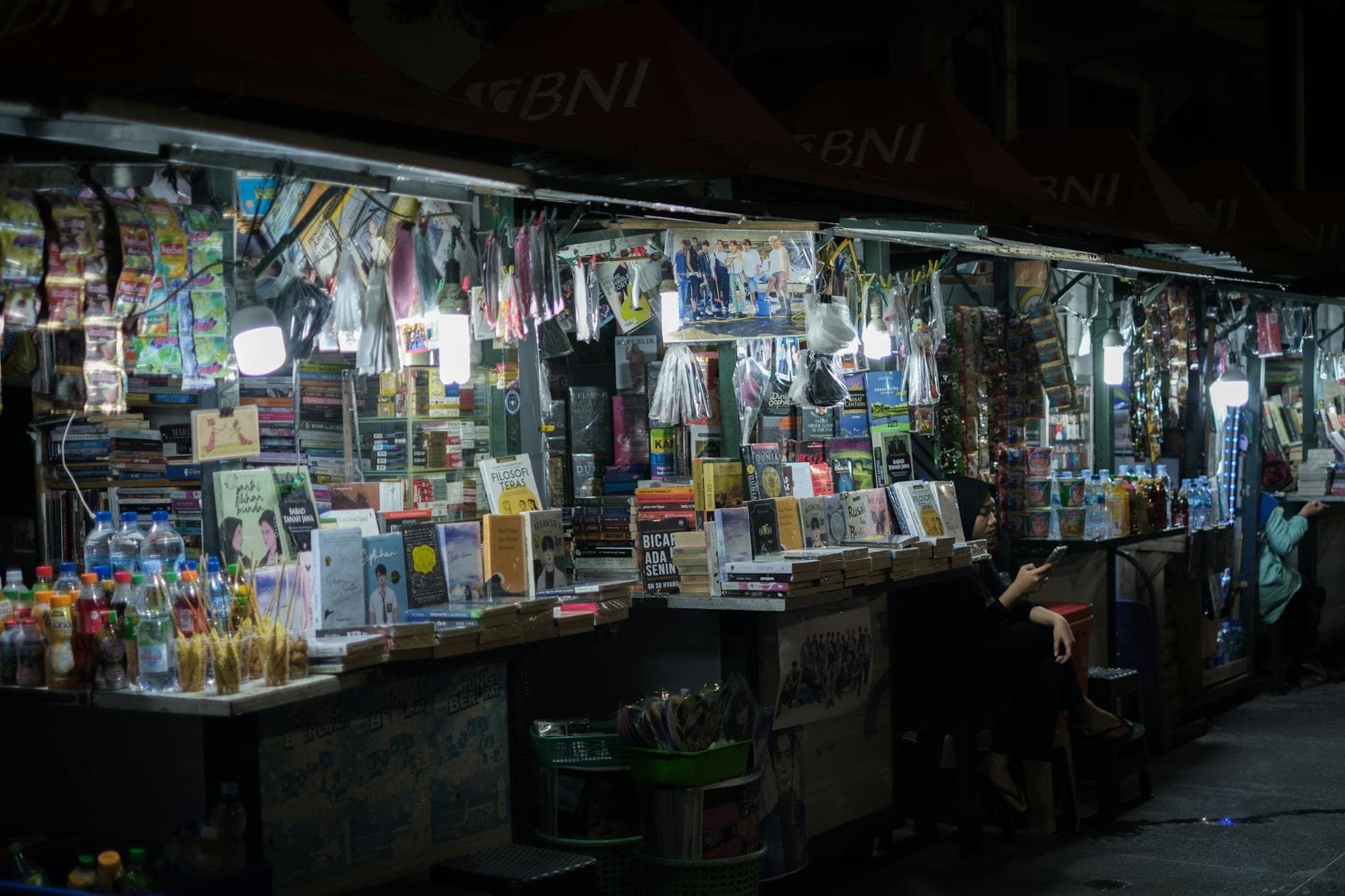 Bustling street market at night featuring book and snack stalls, vibrant atmosphere.