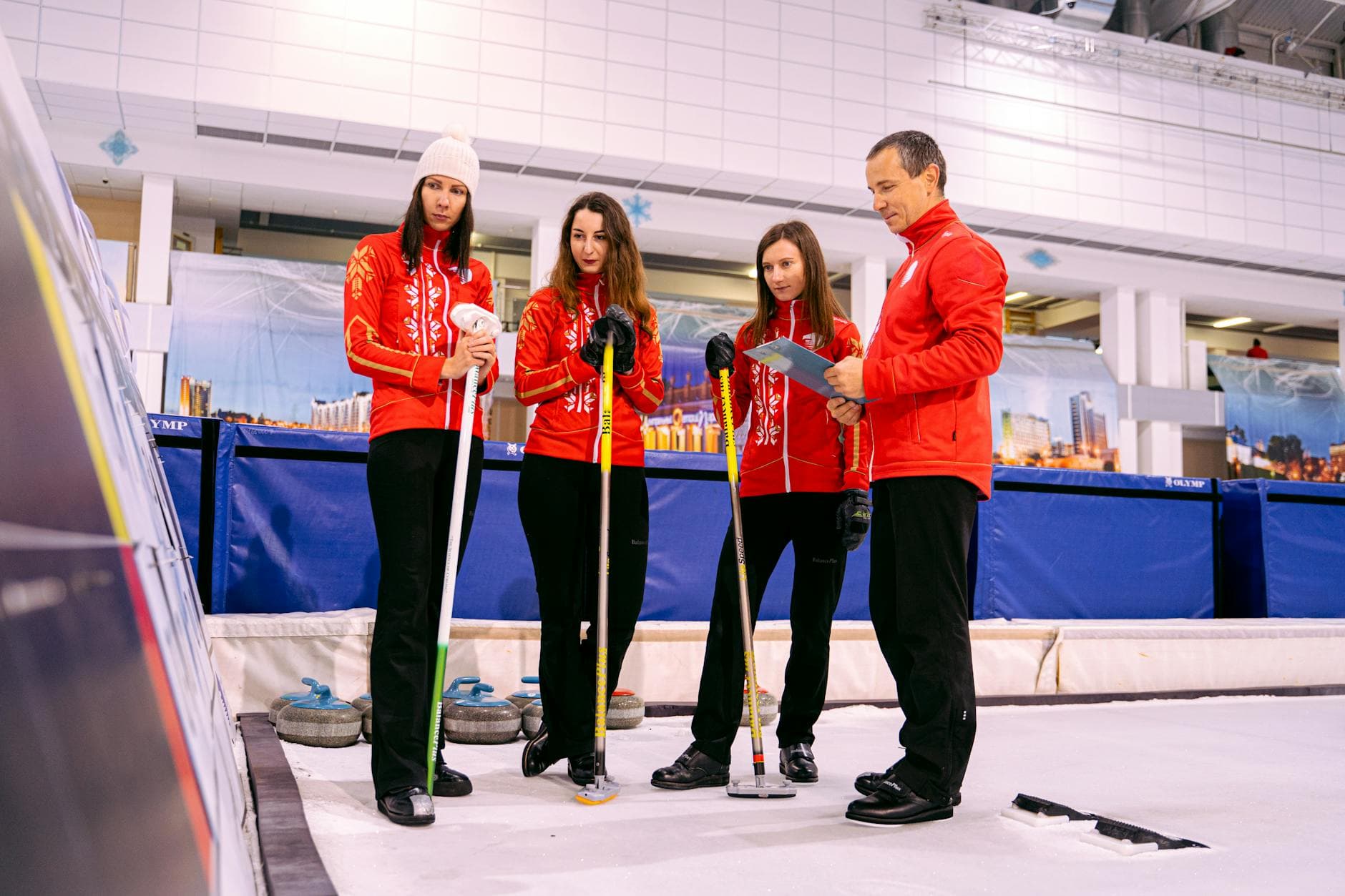 Four curling players in red jackets discussing strategy on an ice rink indoors.