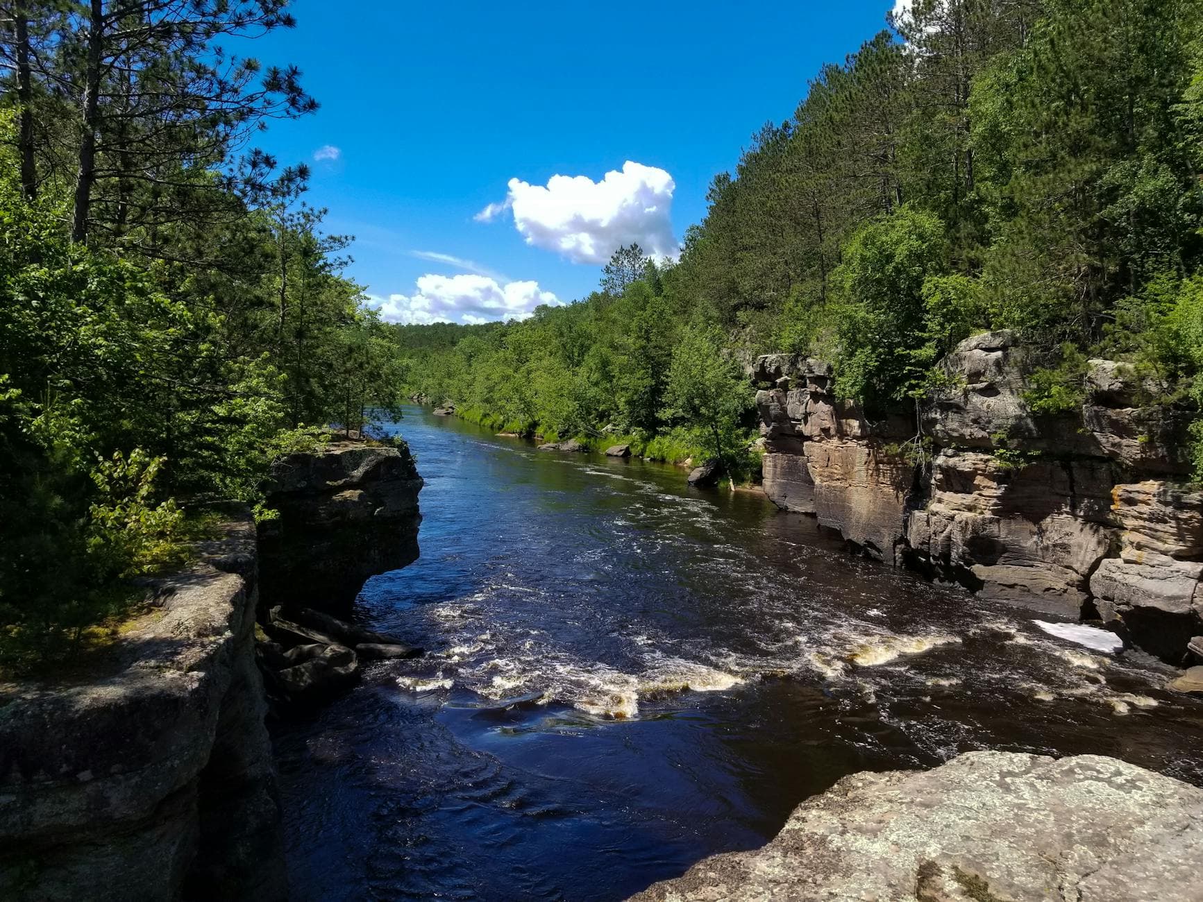 A meandering river bordered by lush greenery and rocky cliffs under a bright blue sky.