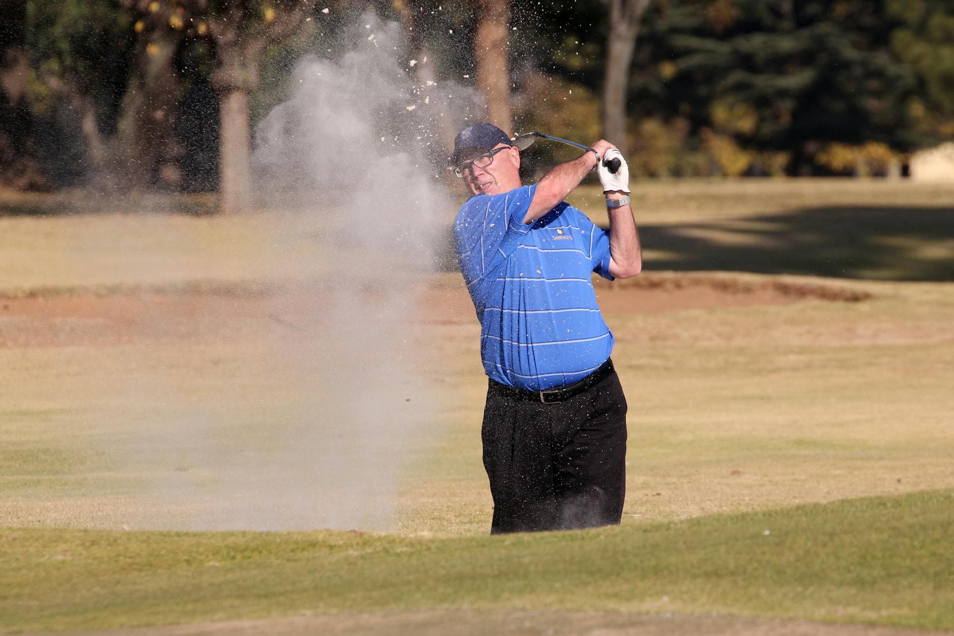 Male golfer executing a sand shot on a golf course under clear blue skies.