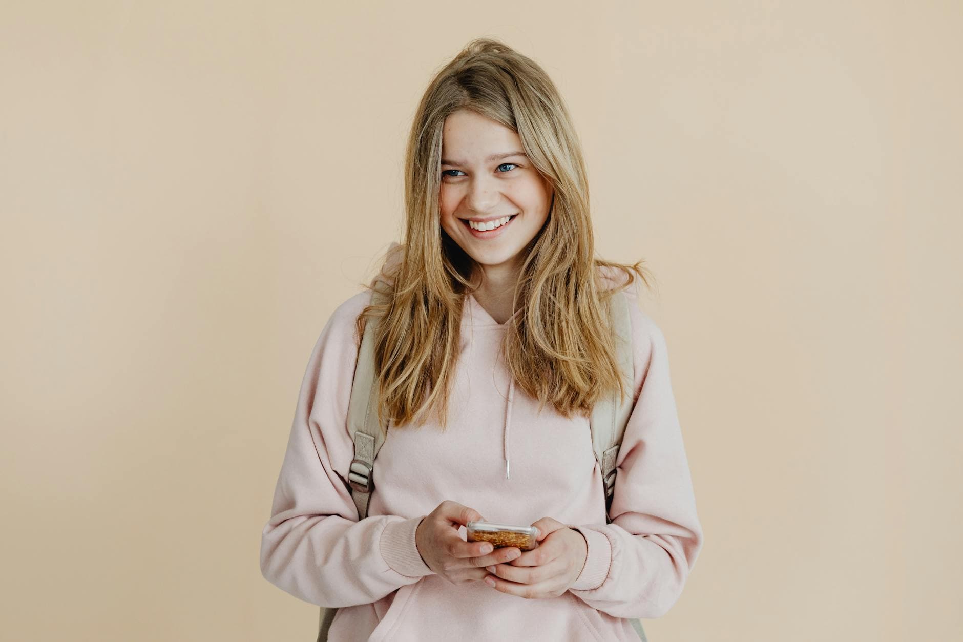 A cheerful teenager with blonde hair wearing a pink sweater and using a smartphone indoors.