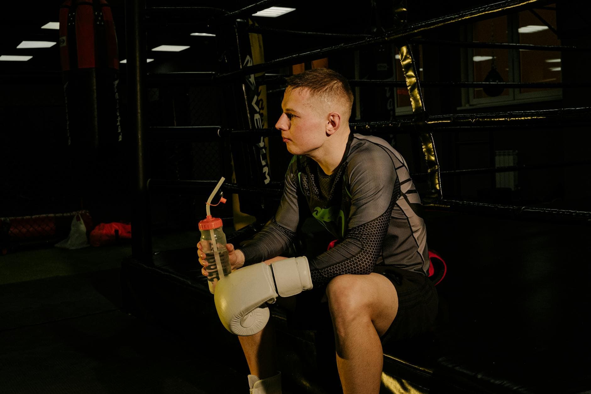 Male boxer sitting in a boxing ring, wearing gloves, and holding a water bottle.