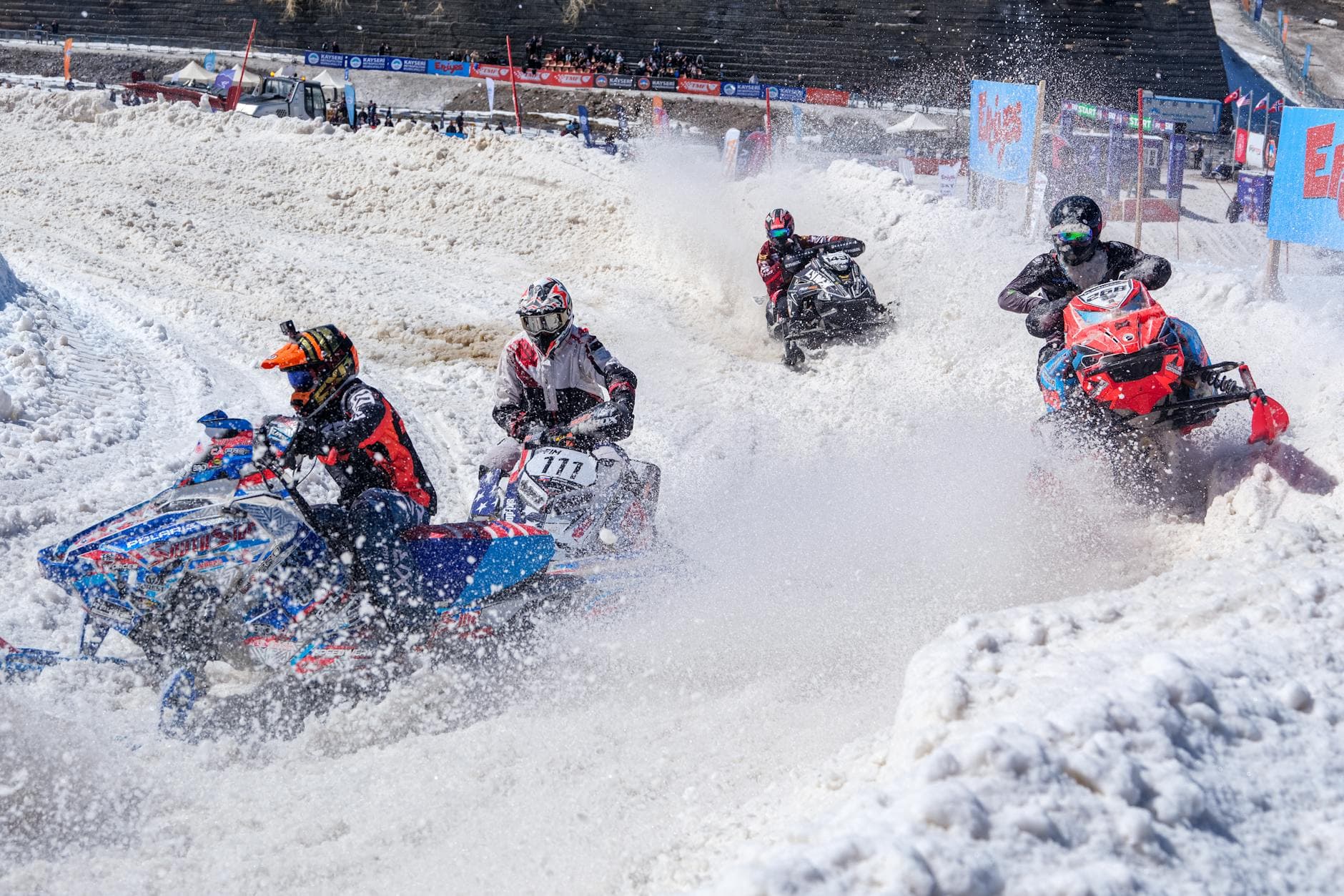 Action-packed snowmobile race on a snowy track with riders making sharp turns amidst flying snow.