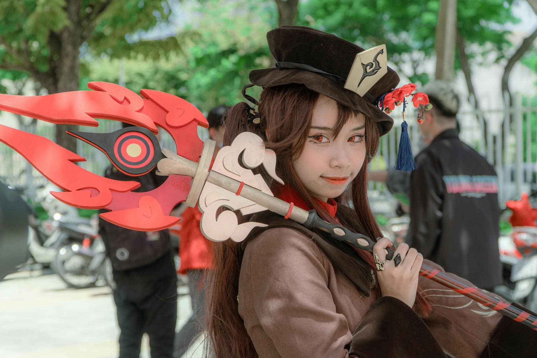 A female cosplayer dressed as Hu Tao from Genshin Impact poses outdoors with a detailed costume and weapon.