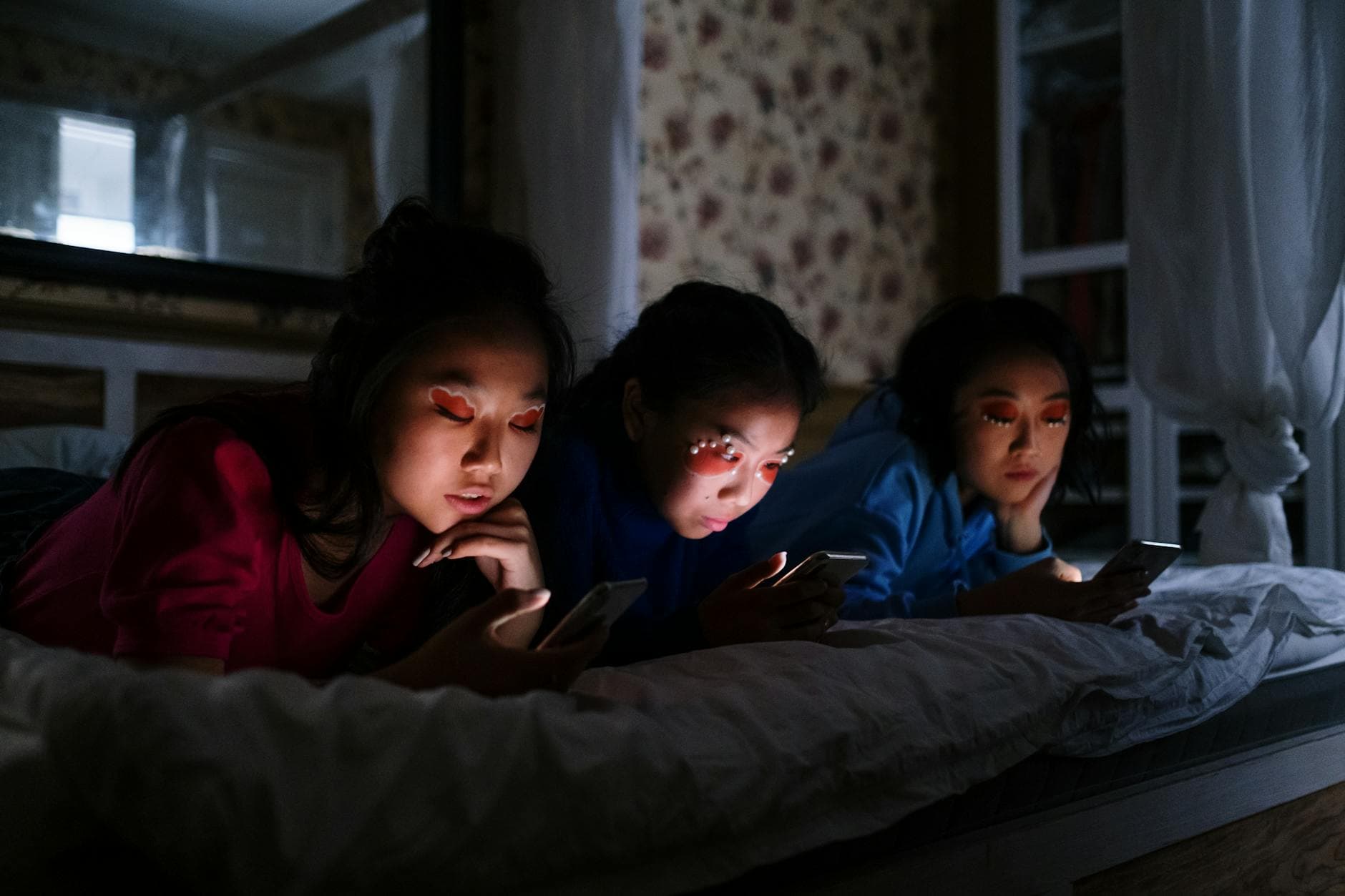 Three teenagers lying on a bed using smartphones at night in a cozy bedroom.
