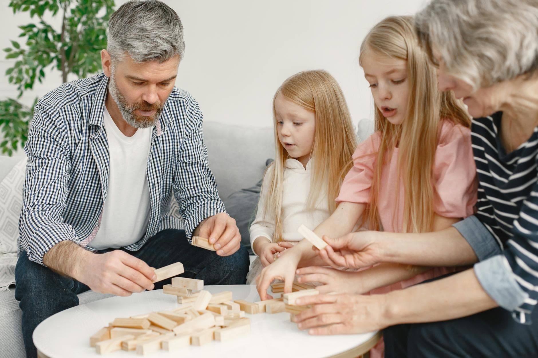Four family members of different ages engage with toy blocks on a table indoors.
