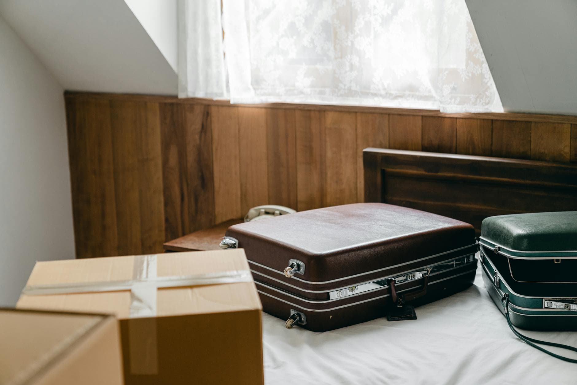 From above of brown and green vintage suitcases and packed carton boxes placed on bed with sheet in light room with sloping roof