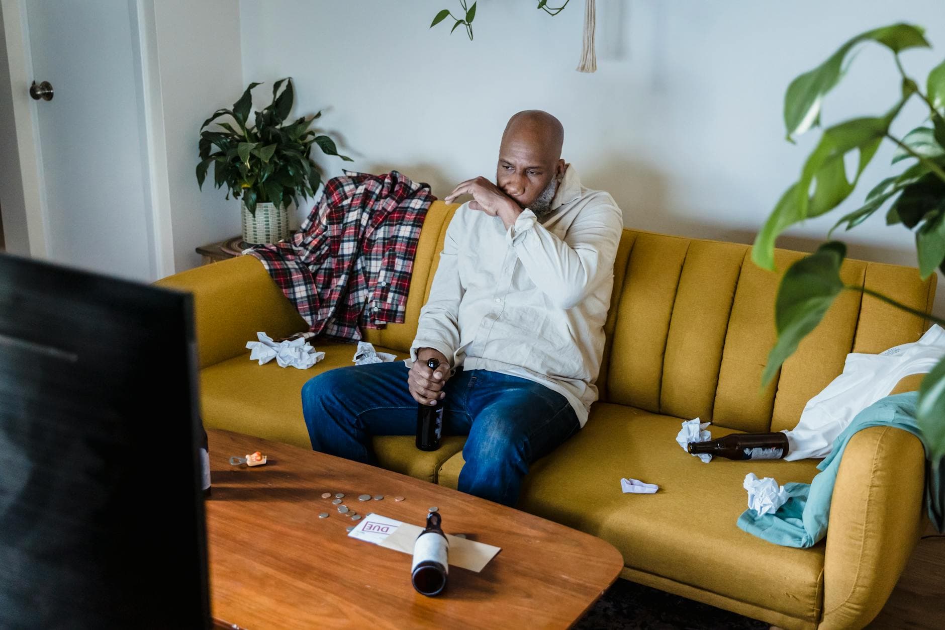 An adult man sitting on a sofa looking distressed, surrounded by financial documents and mess.
