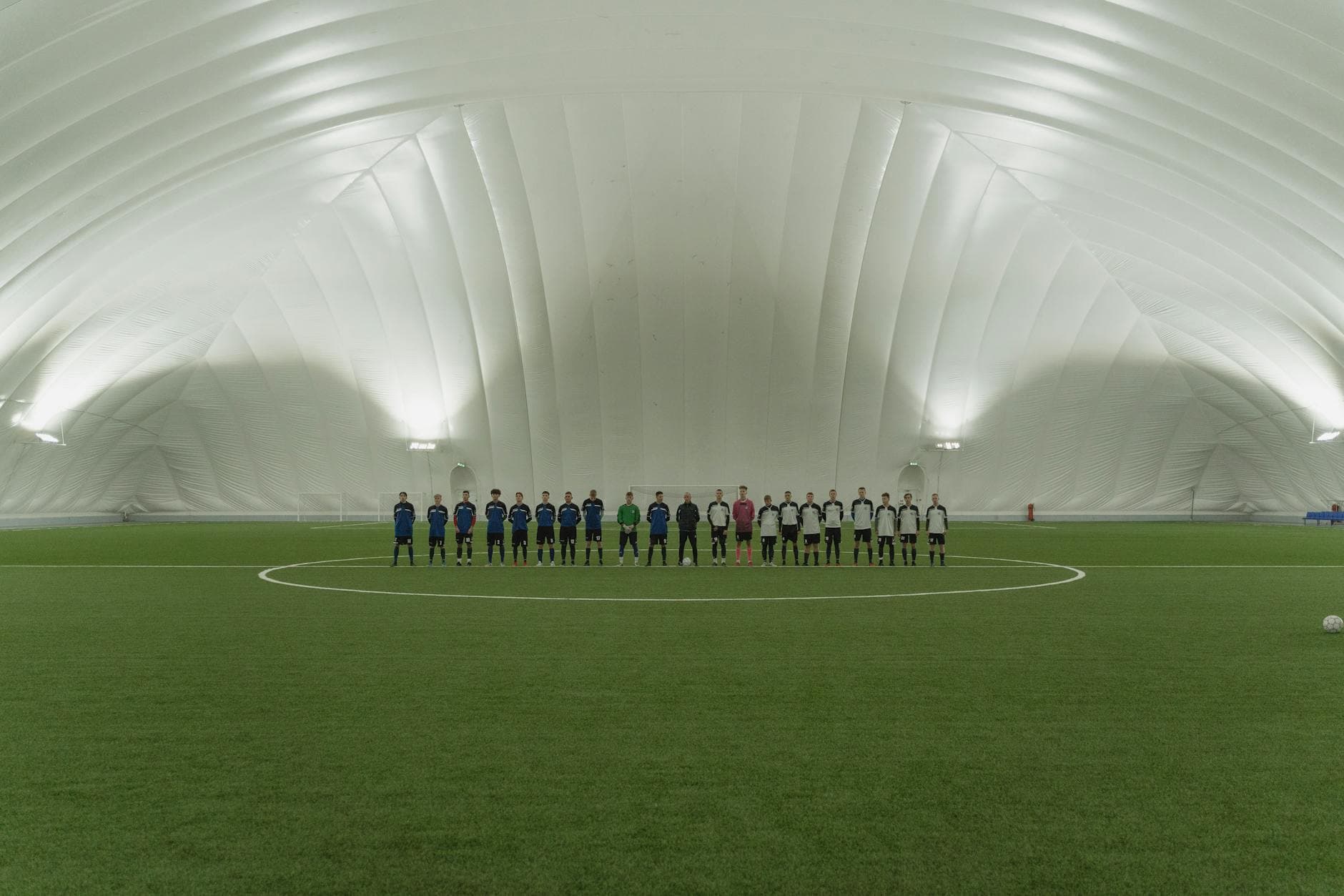 Two soccer teams lined up on an indoor artificial turf field under dome lighting.