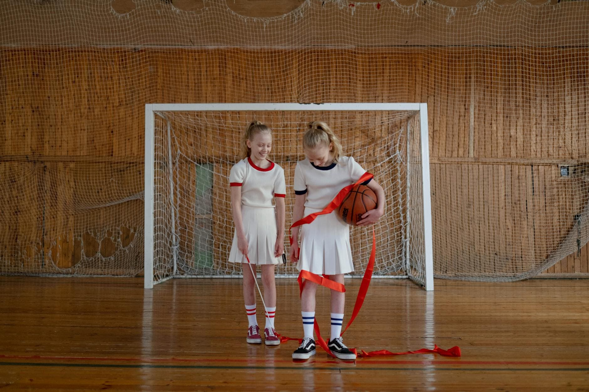 Two young girls standing in a gym celebrating with a basketball and ribbon.