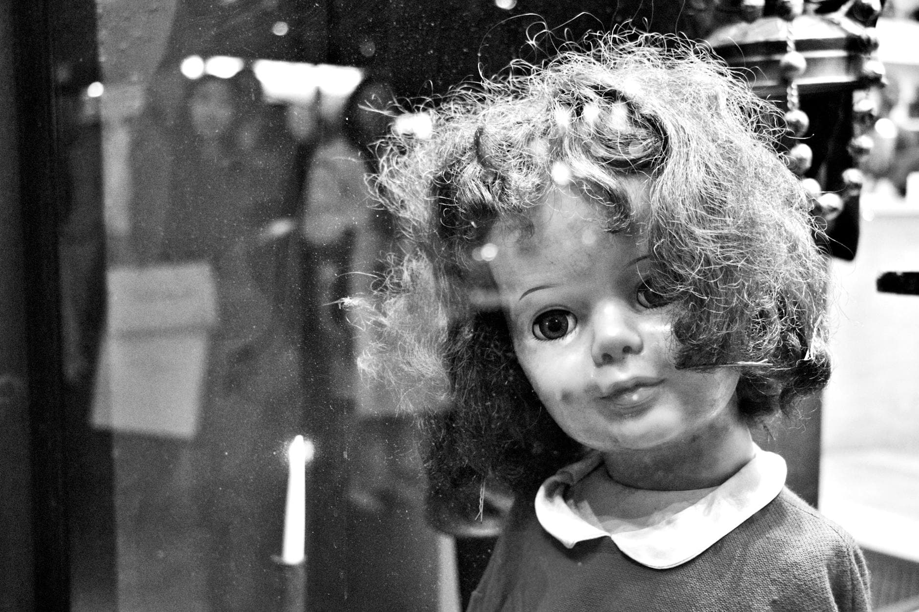 Black and white image of a vintage doll with a creepy expression seen through glass.
