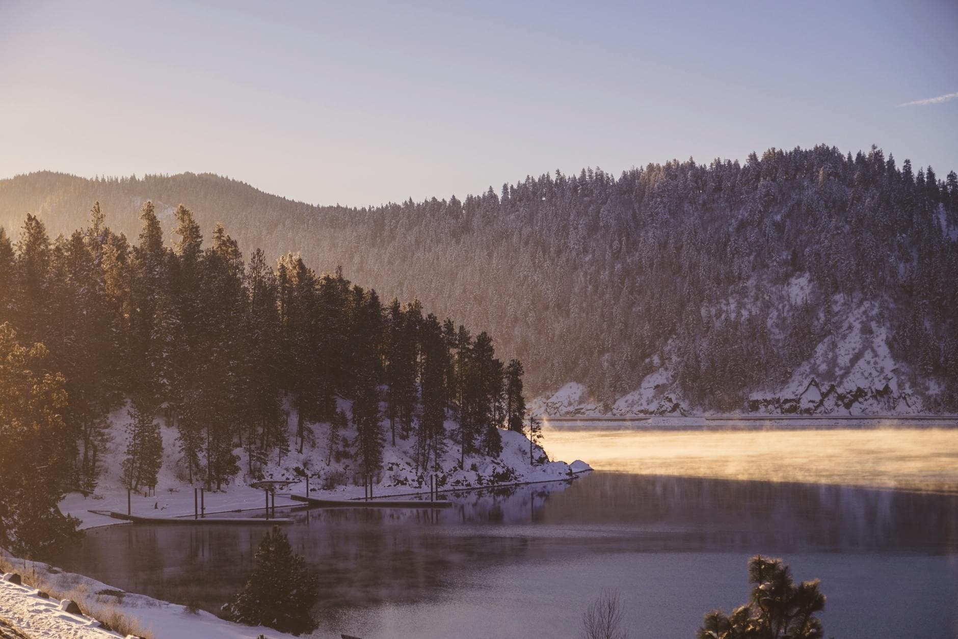 Peaceful winter lake with mist rising over snow-covered mountains at sunrise.
