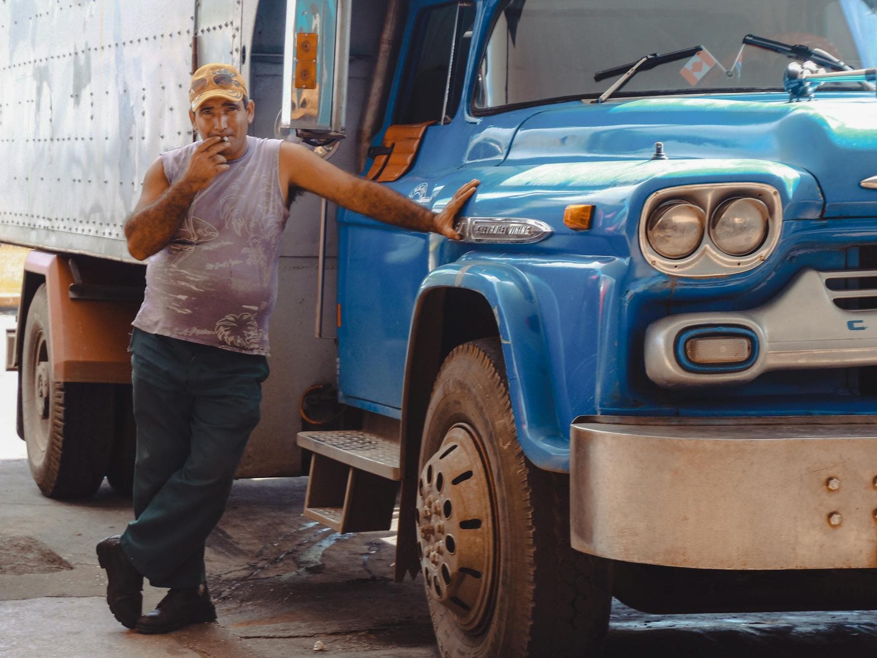 A middle-aged man smokes a cigarette while leaning on a blue vintage truck outdoors.