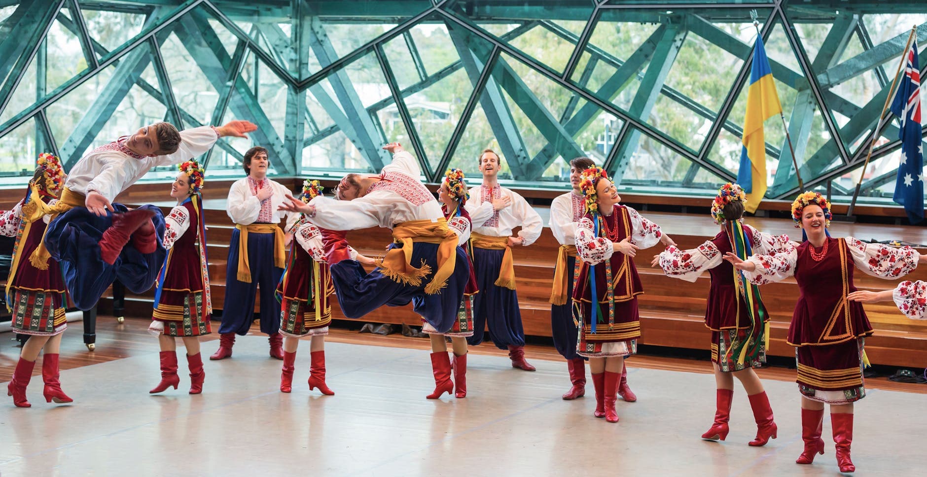 Group performing vibrant traditional folk dance indoors with flags visible.