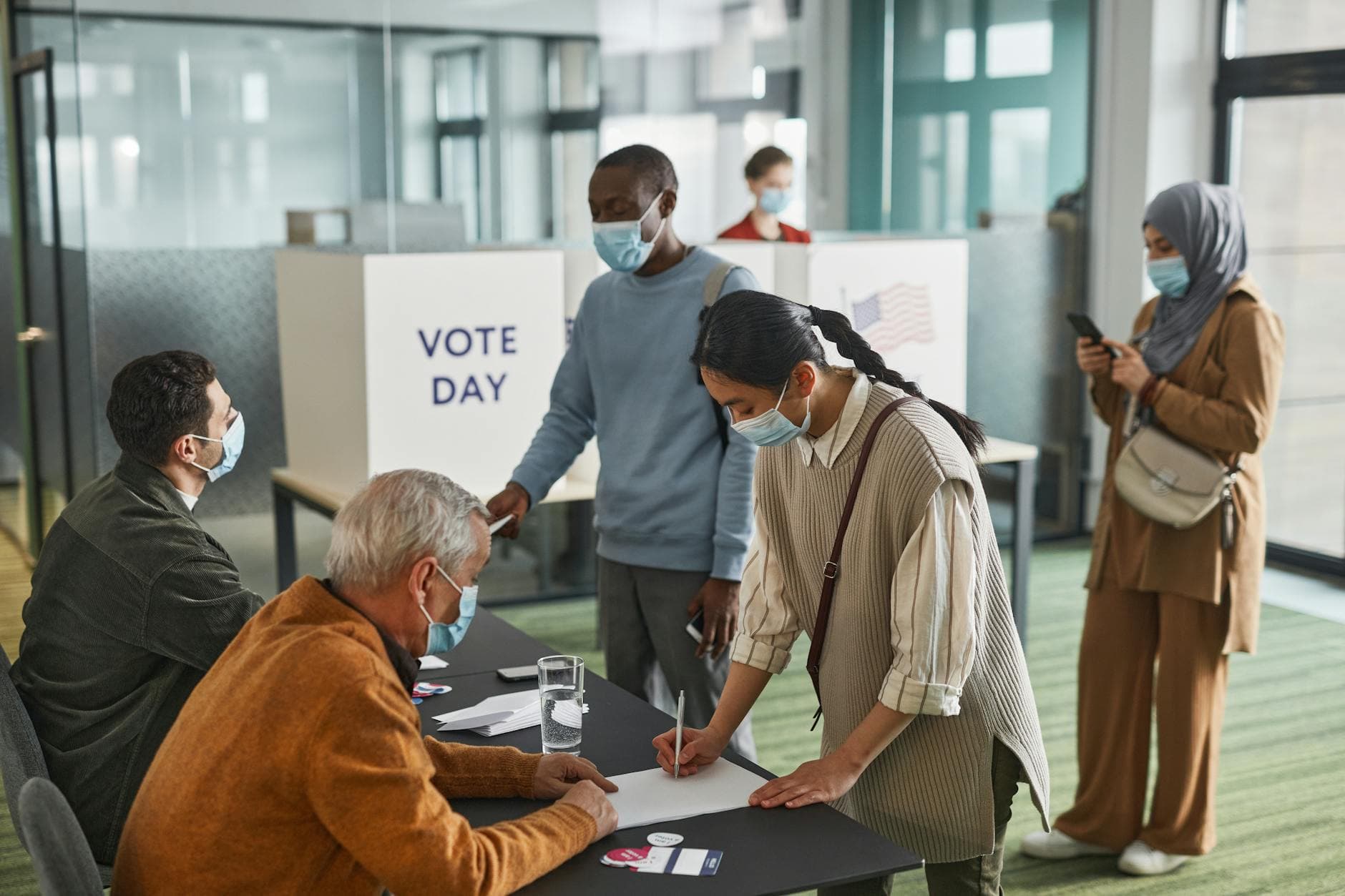 A diverse group of adults casting votes indoors on election day, maintaining safety protocols.