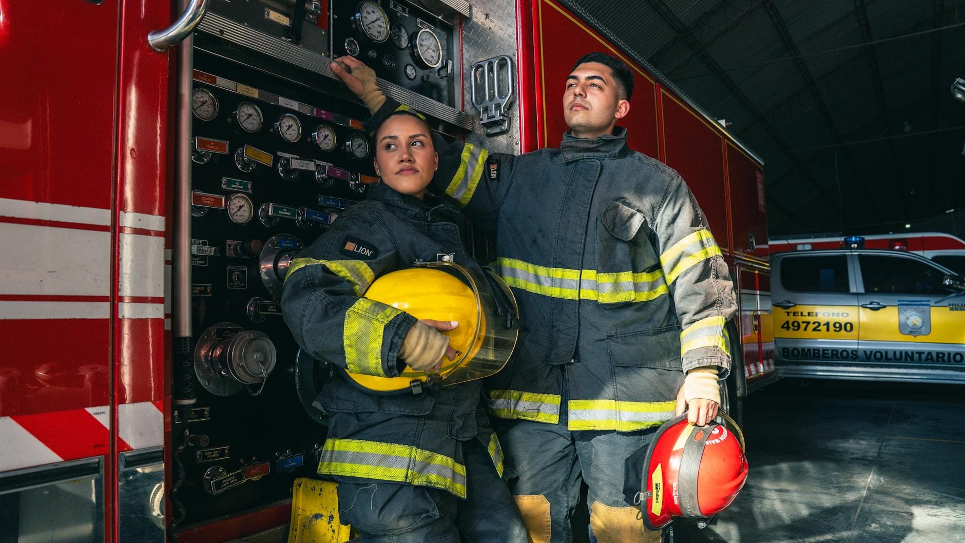 Two firefighters in full gear stand by a firetruck inside a station.