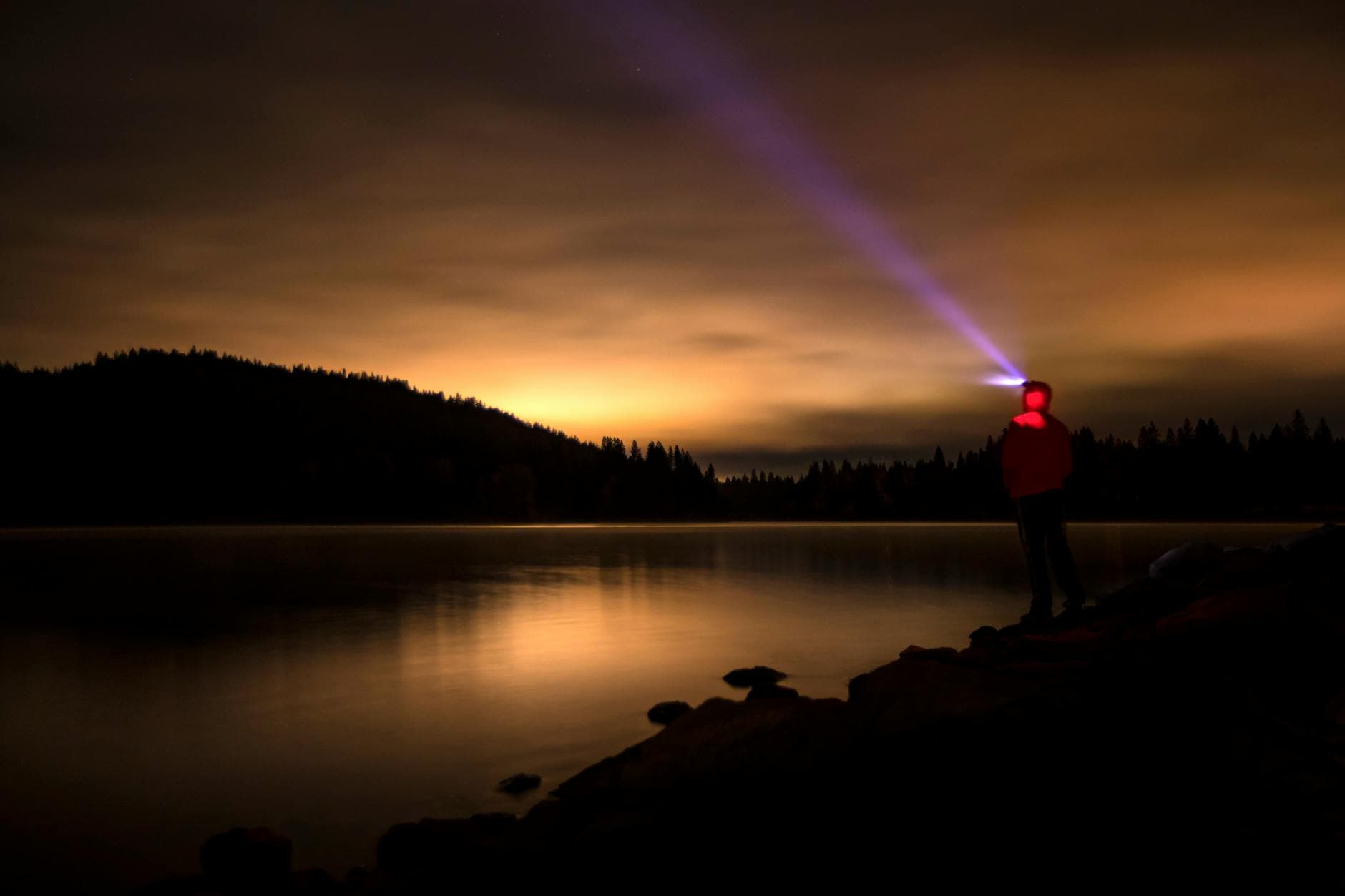 A solitary figure with a headlamp gazes over a calm lake under a vibrant night sky.