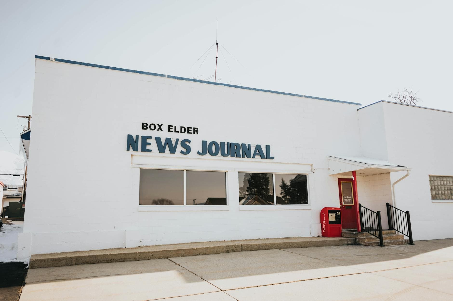 Exterior view of Box Elder News Journal building with red door and sign.