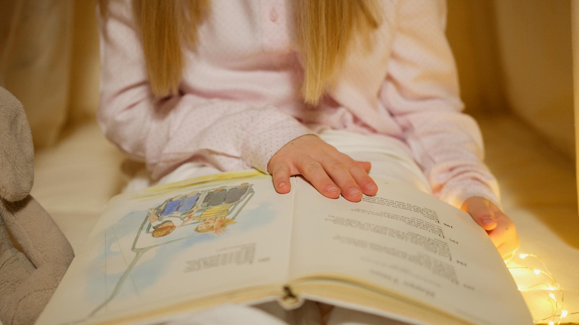 A young girl in pajamas reading a storybook in a warm, cozy bedroom setting.