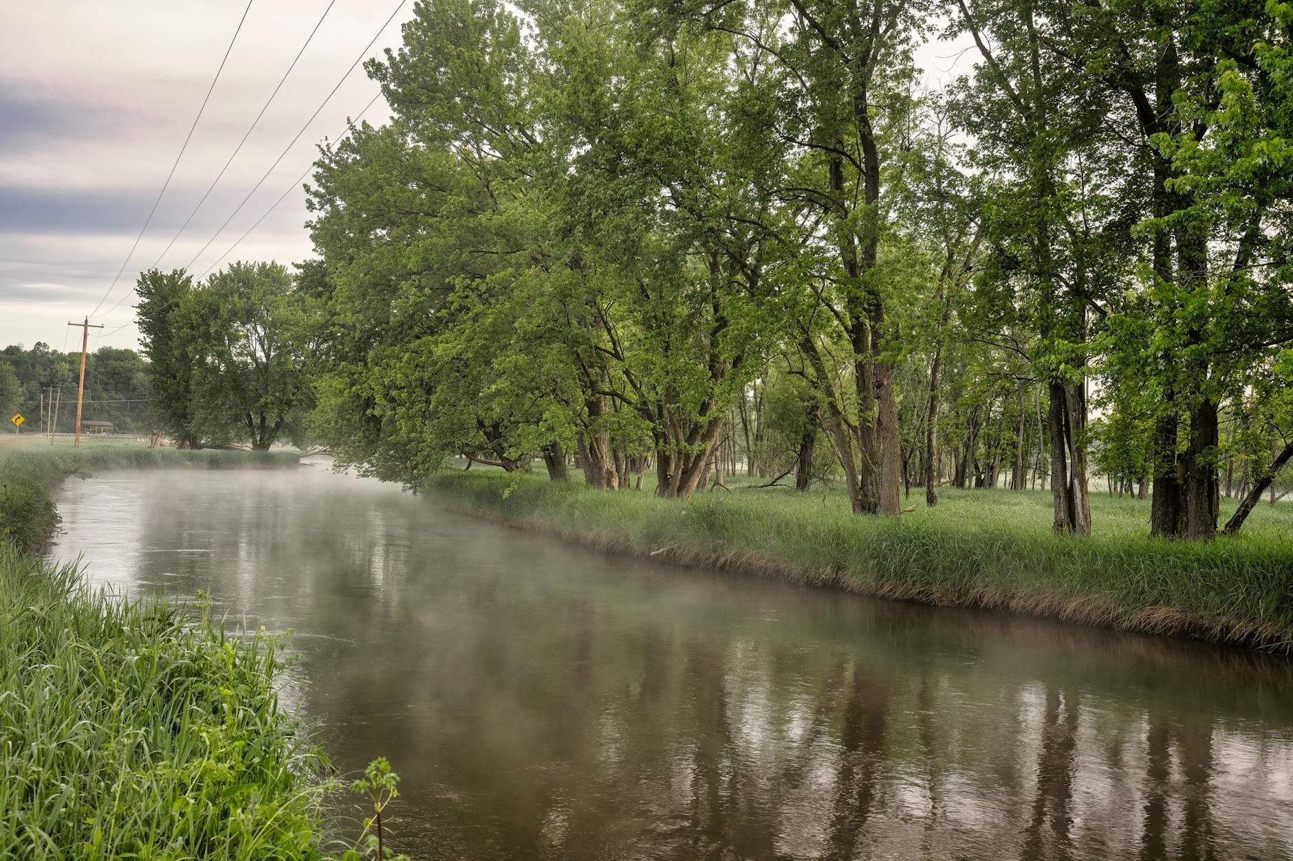 Serene river with mist rising, surrounded by lush trees and electricity lines in a rural setting.