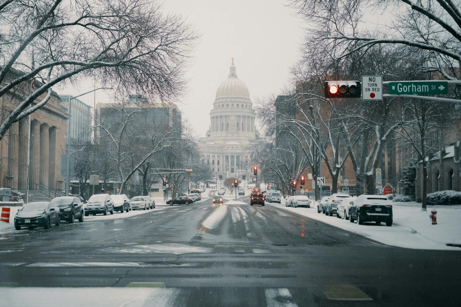 A serene winter view of the Wisconsin State Capitol under snowfall with snow-covered streets and cars.