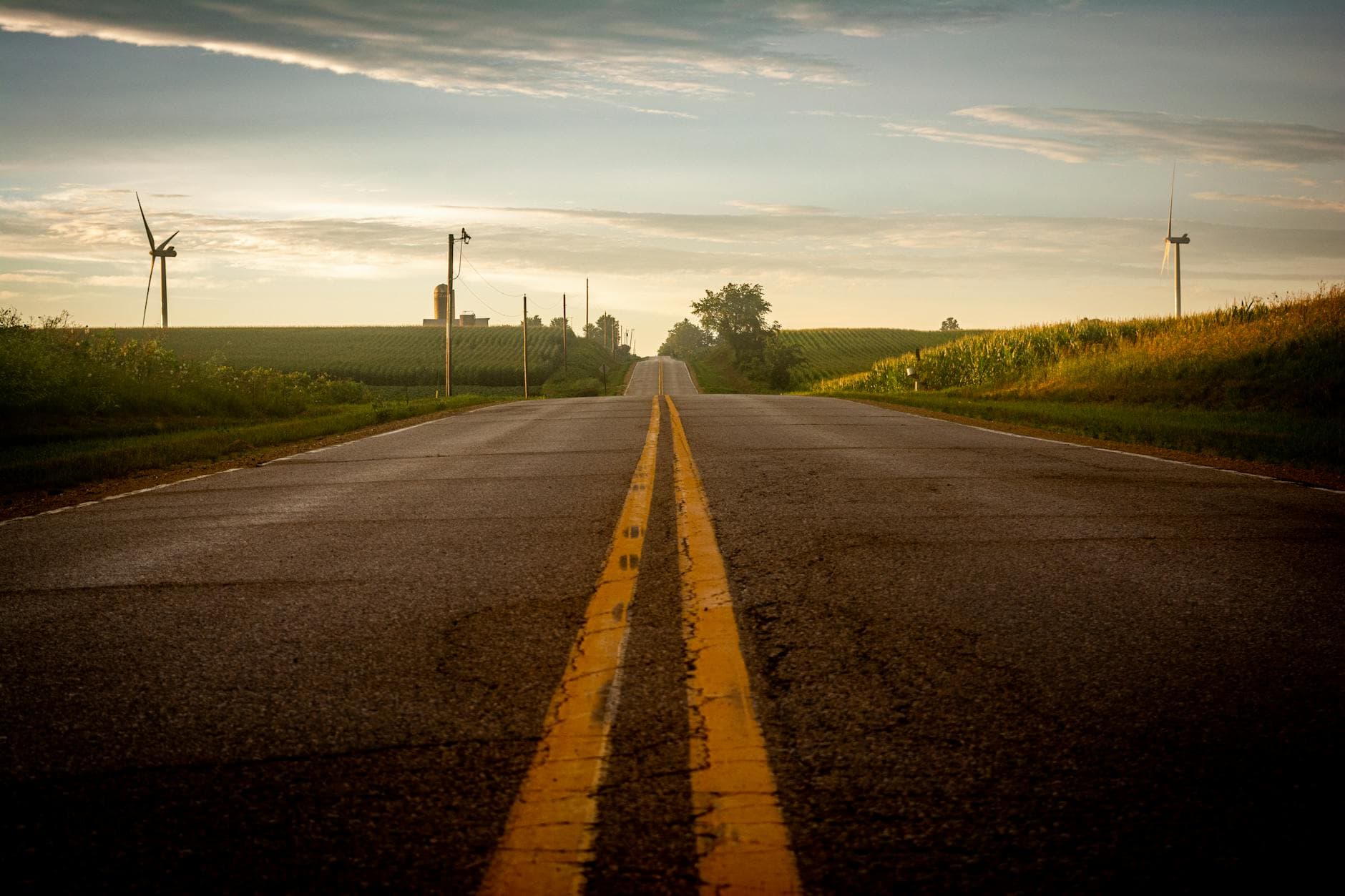 A scenic rural road flanked by wind turbines and fields under a summer sky.