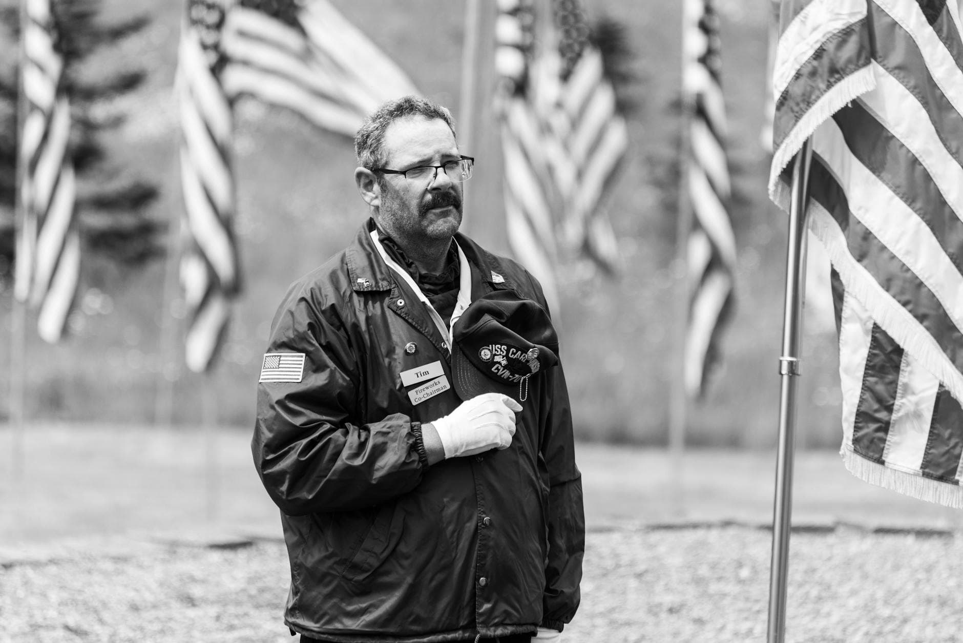 A veteran holding a hat stands solemnly among flags at a military memorial in black and white.
