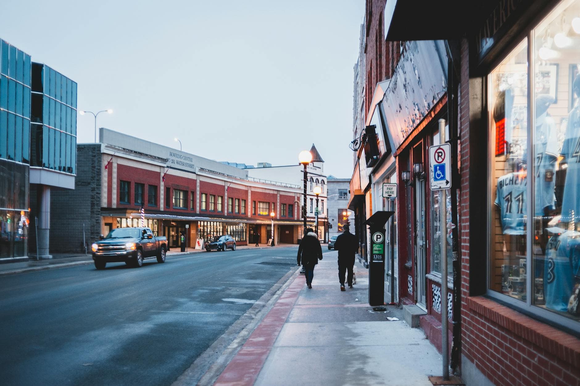 A lively urban street with storefronts, pedestrians, and vehicles during the day, evoking a bustling atmosphere.