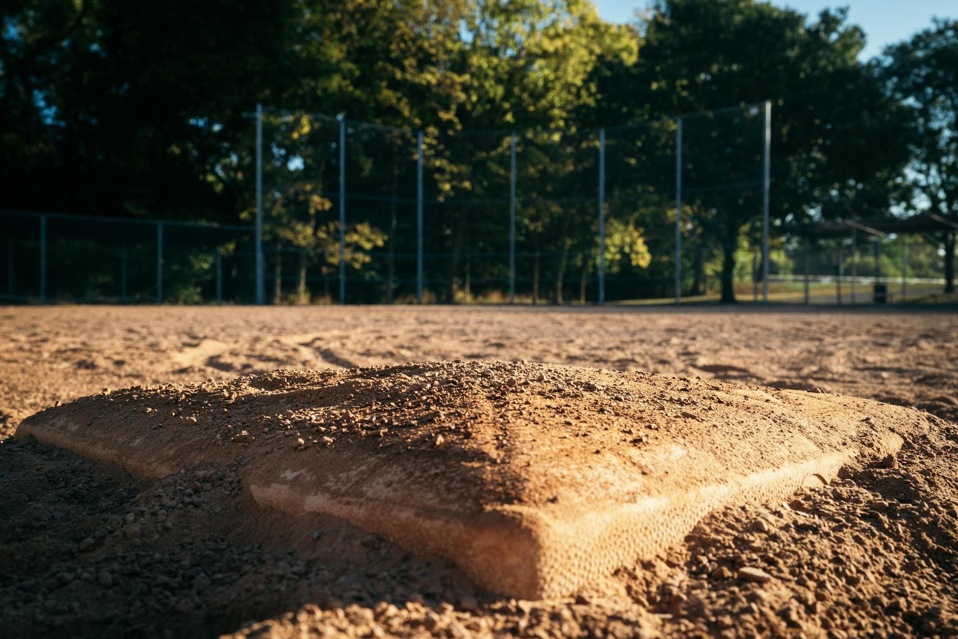 Close-up of a baseball base on an empty field with trees and fence in the background.