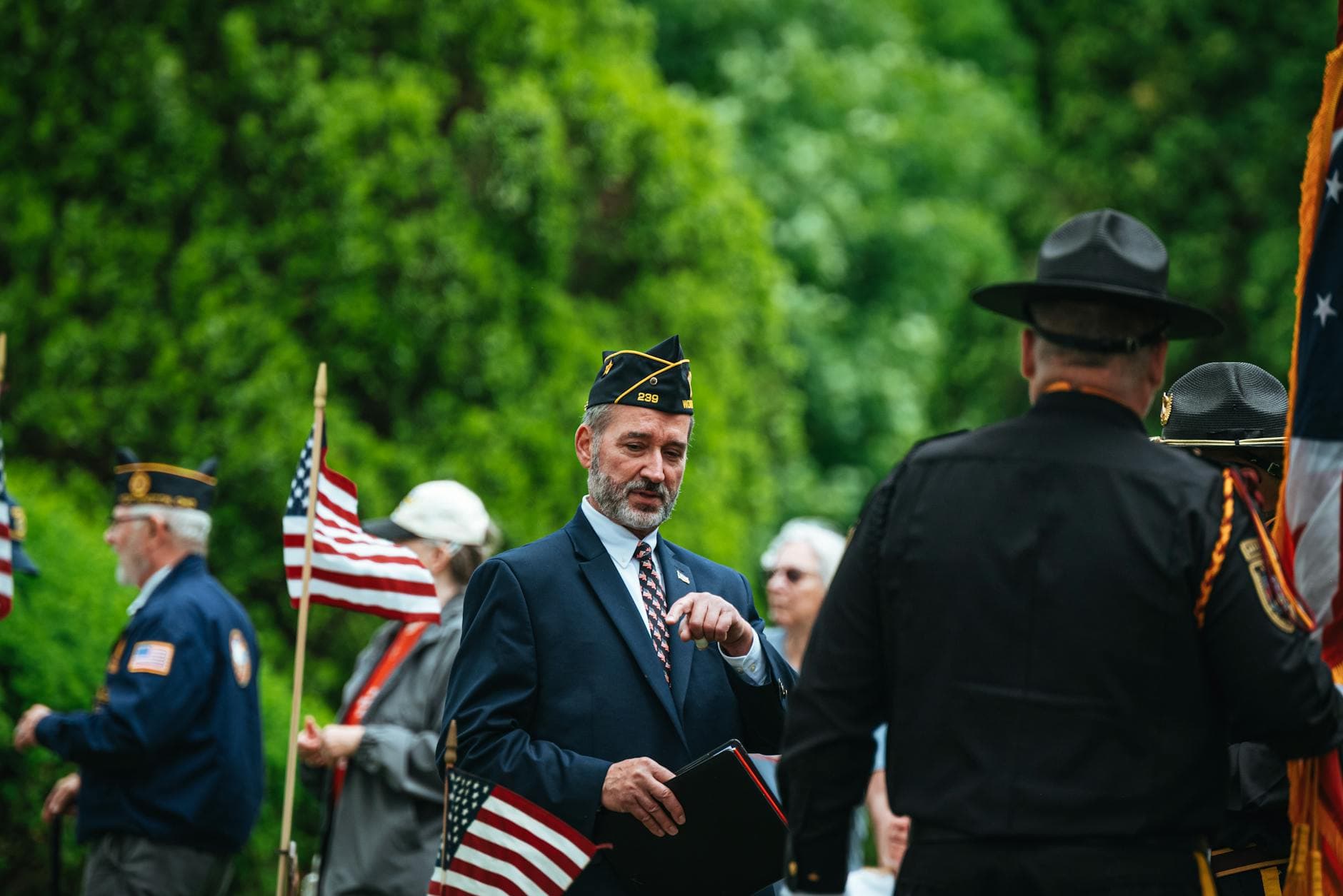 Veterans gathered in formal attire holding American flags during a memorial ceremony outdoors.