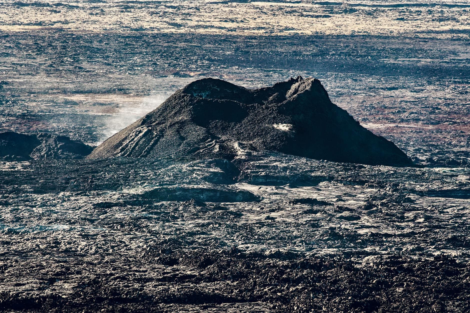 An expansive aerial view of a rugged lava field with a volcano at its center.