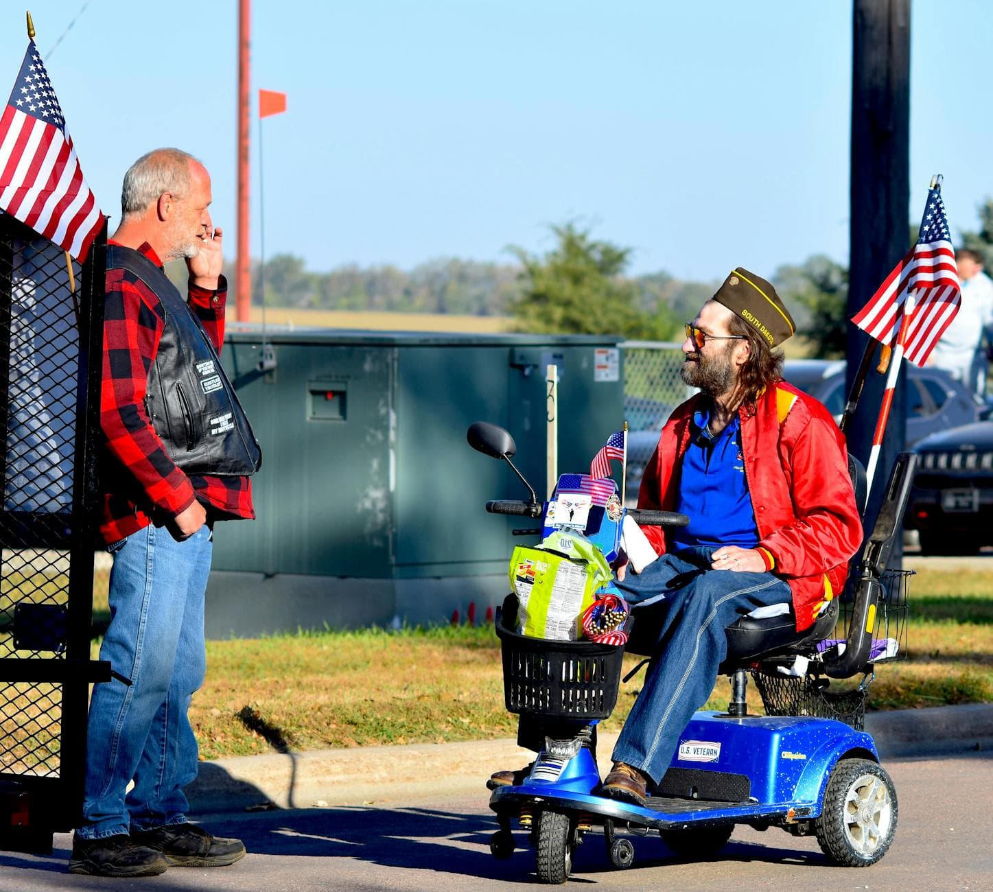 Two veterans converse outdoors beside American flags, symbolizing patriotism and camaraderie.