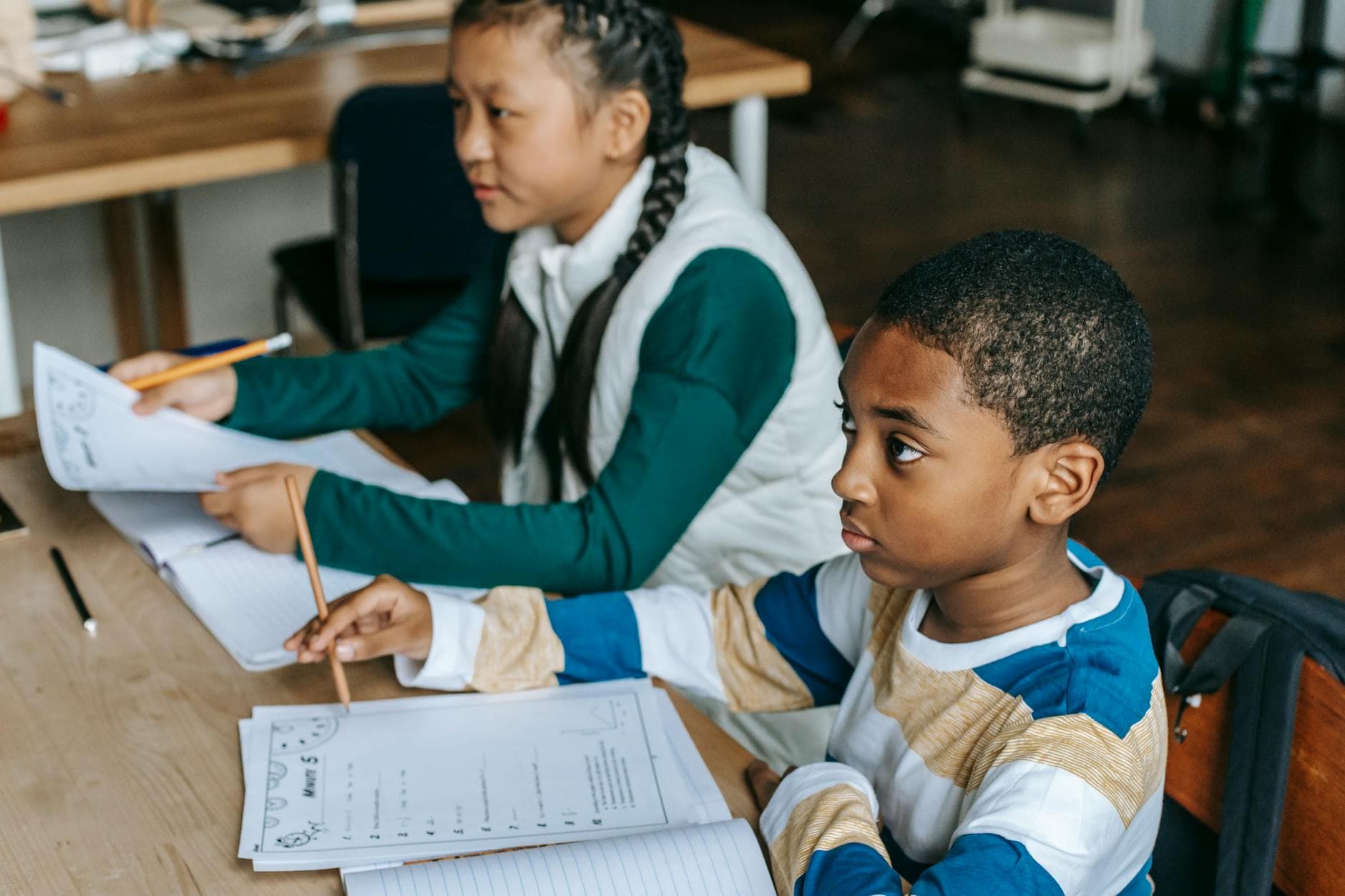 Side view diligent multiracial elementary pupils wearing casual clothes sitting at desk with notebooks and looking away