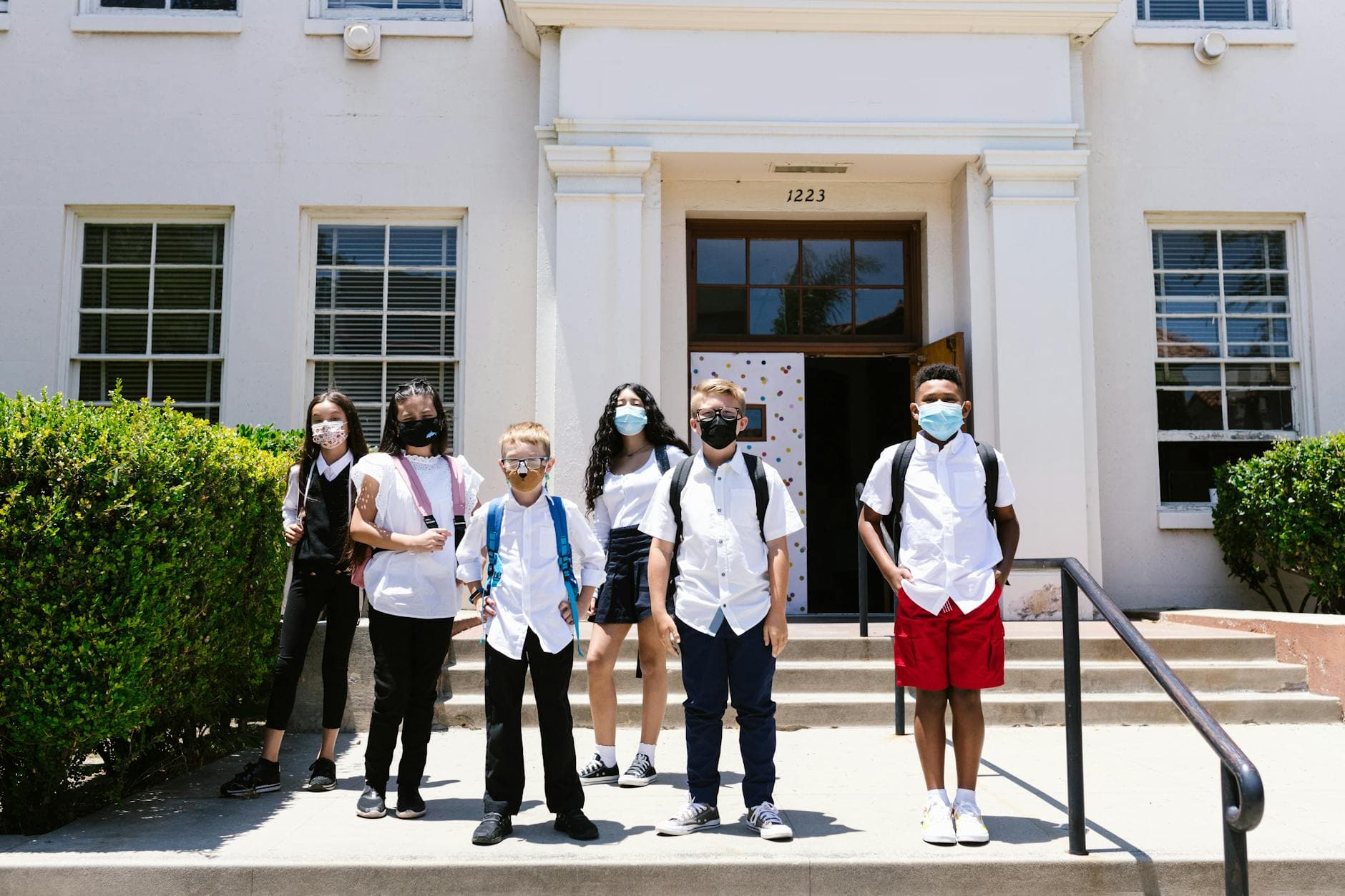 Students in masks and uniforms standing outside a school building on a sunny day.